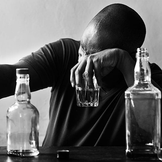 sober living | African American man drinking alcohol with bottles around depressed looking tired