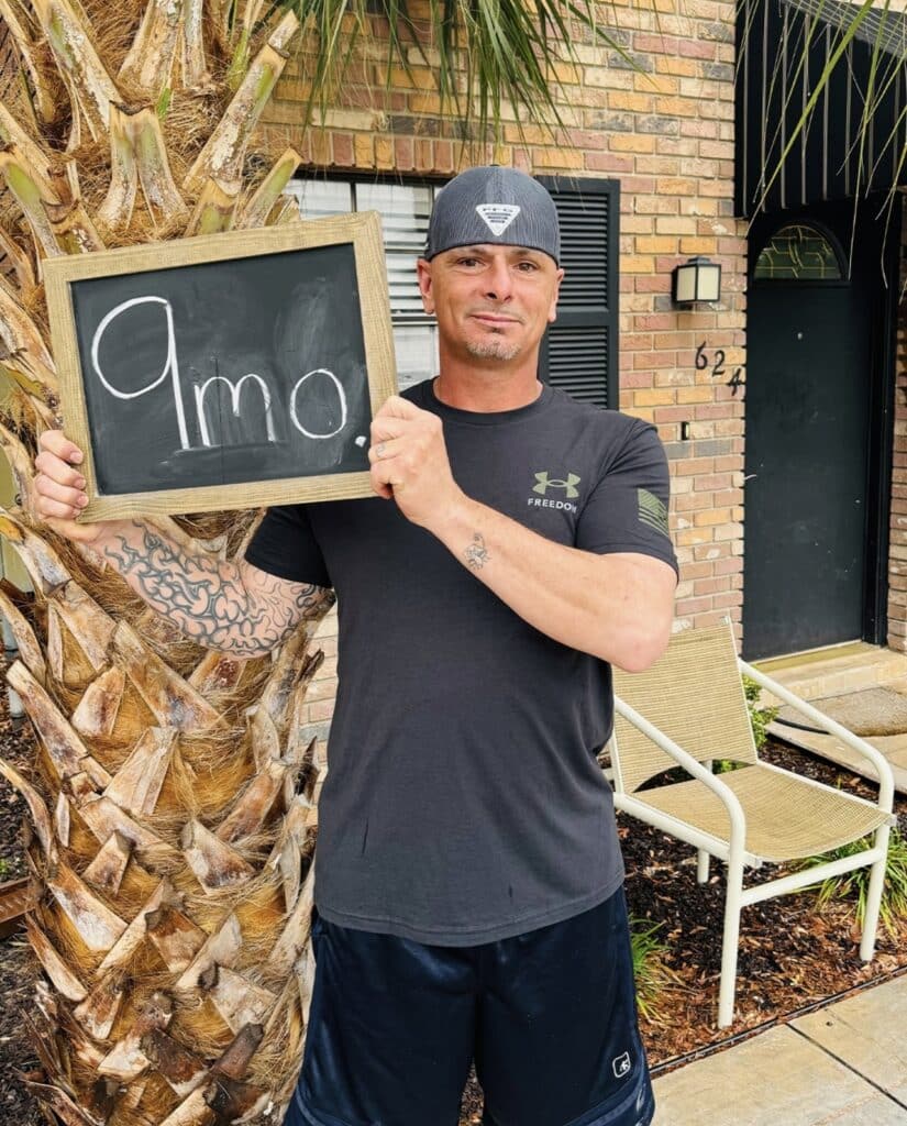 man holding 9 months sober sign in front palm tree and building